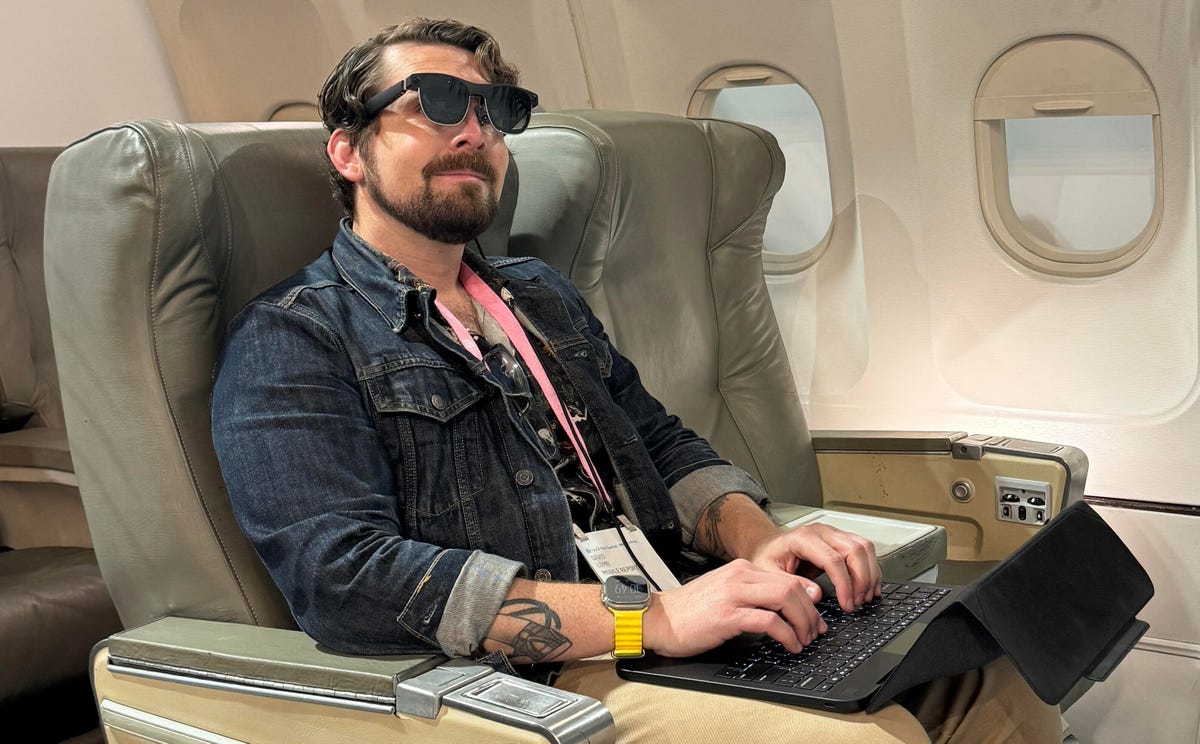 A man wears AR glasses connected to a laptop (without a display) while sitting in the seat of a mock-up airplane.